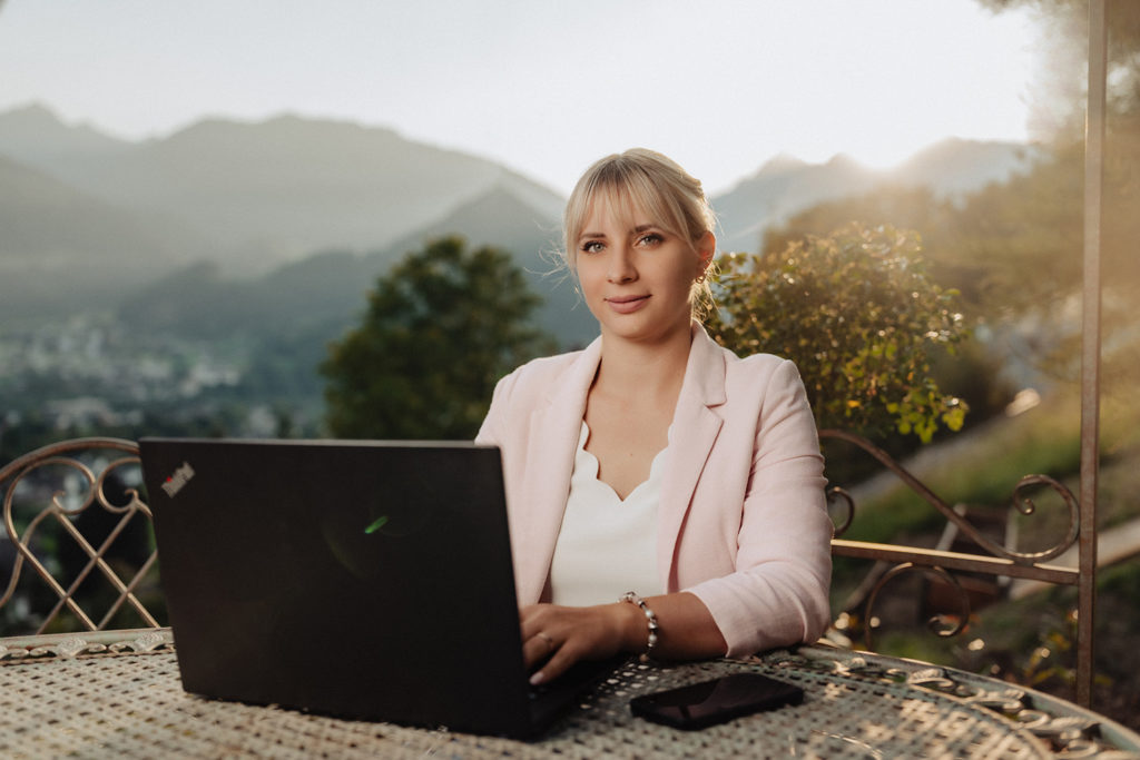 Florine Fürstler, junge blonde Frau vor Laptop mit Sonnenuntergang im Hintergrund und Blick auf die Berge